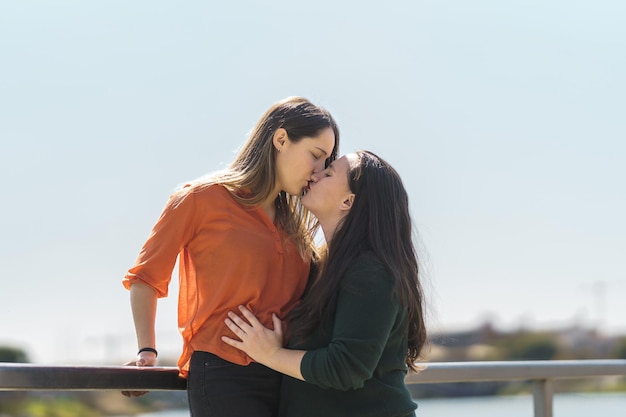 Lesbian couple kissing while one of them is climbing on a railing next to river