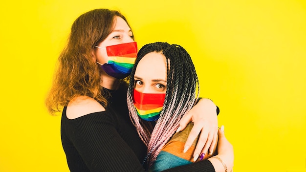 Lesbian couple in rainbow masks embracing in studio side view\
of lgbt women in colorful rainbow masks hugging on yellow\
background in studio