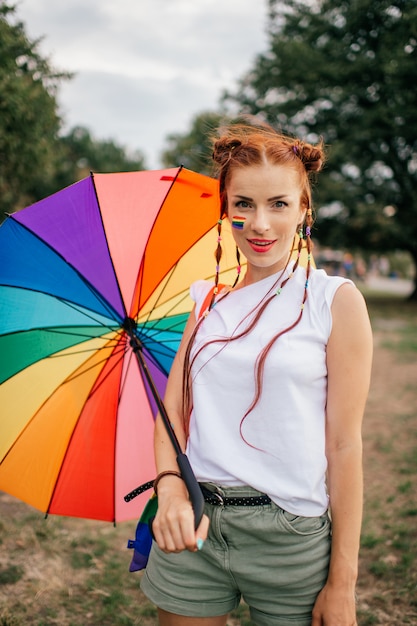 Lesbisch rood haarmeisje met vlechten, lgbt-vlag op haar gezicht, kleurrijke zak en paraplu openlucht stellen