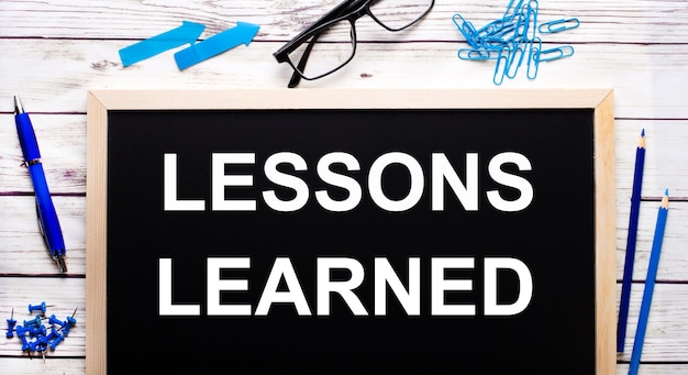 LESSONS LEARNED written on a black note-board next to blue paper clips, pencils and a pen.