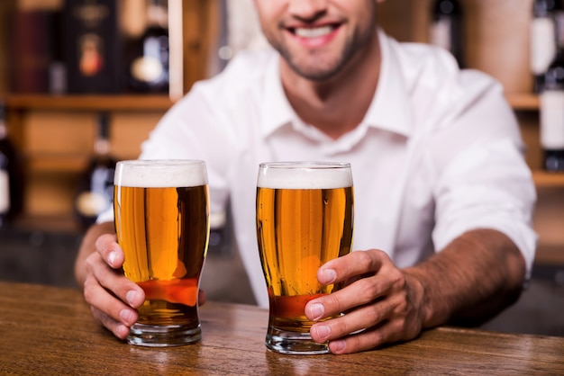 Lest je dorst! Close-up van knappe jonge mannelijke barman in wit overhemd die glazen met bier uitrekt en glimlacht terwijl hij aan de toog staat