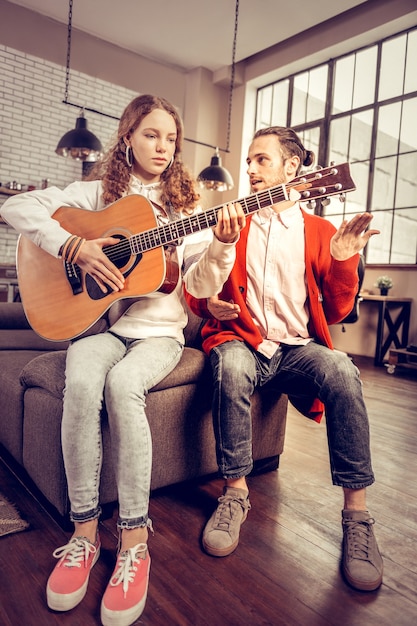 Lestijd. Jonge ervaren gitaarleraar met rood vest die tienermeisje gitaar leert spelen