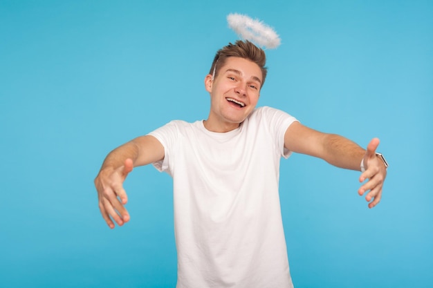 Photo let me embrace you. angelic kind-hearted man with halo above head outstretching hands to hug and looking at camera with childish joyful expression. indoor studio shot isolated on blue background