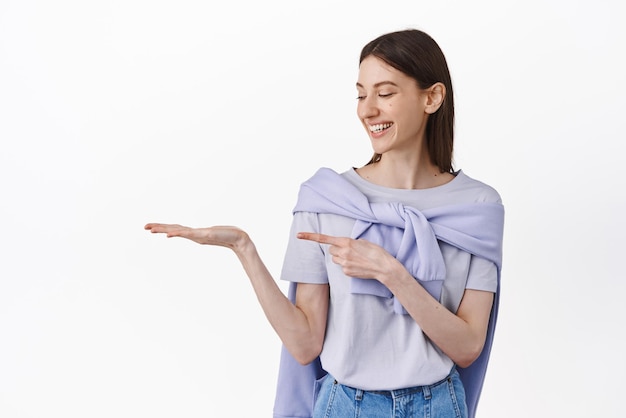 Let me introduce you this Pretty young woman looking at her open hand holding logo product pointing at empty space on her palm showing something white background