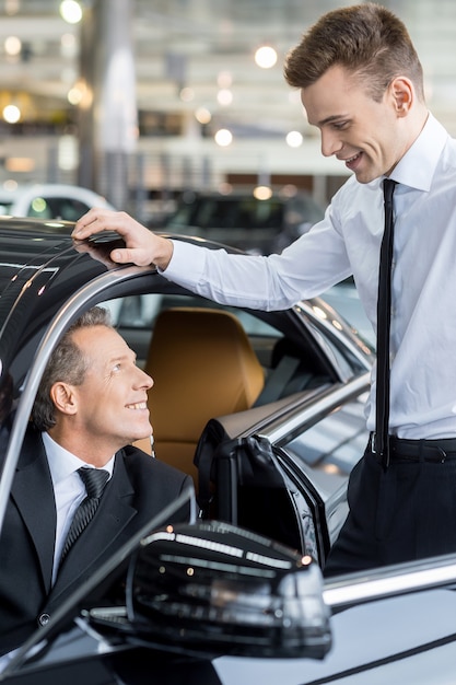 Let me show you all the features. Confident mature man ion formalwear sitting at the front seat of the car and talking to the car salesman standing near him