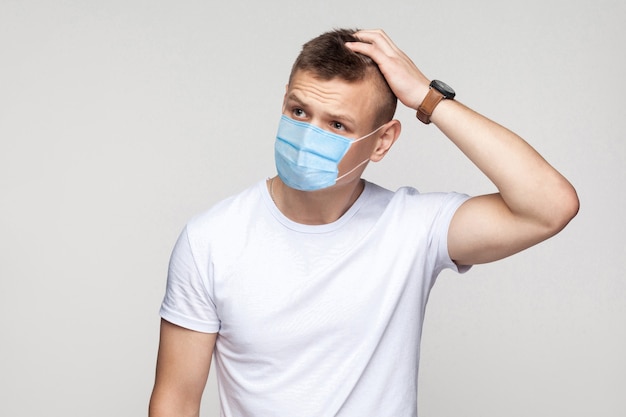 Let me think. Portrait of thoughtful young man in white shirt with surgical medical mask standing, scratching his head and thinking what to do. indoor studio shot, isolated on gray background.