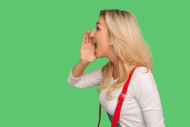 Let op, advertentie. Zijaanzicht van volwassen blonde vrouw in stijlvolle denim overall die luid, hysterisch schreeuwend bericht schreeuwt, idee aankondigend. indoor studio-opname geïsoleerd op groene achtergrond