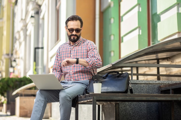 Let op je tijd. Vrolijke volwassen man met behulp van zijn laptop zittend in de stereet