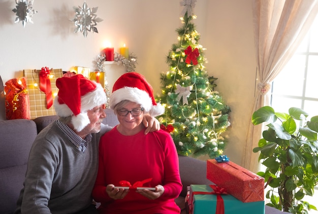 一緒にクリスマスを祝いましょう。贈り物の交換を楽しんでいる美しい老夫婦。彼らはサンタの帽子をかぶっています。背景の美しいクリスマスツリーと家族への贈り物