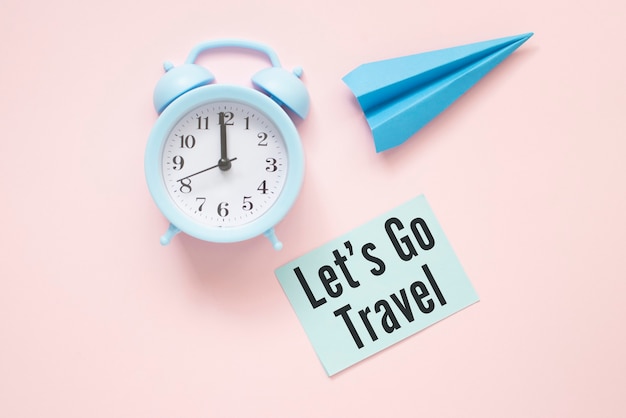Let's Go Travel text on paper with clock and paper airplane on a pink table.