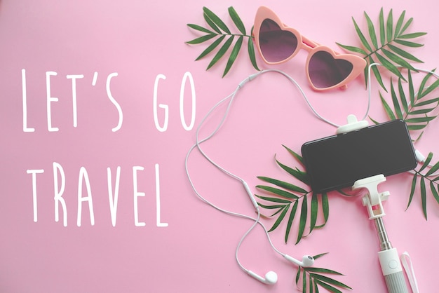 Let's go travel text on stylish pink sunglasses phone on selfie
stick headphones and green palm leaves on pink background summer
vacation flat lay time to travel