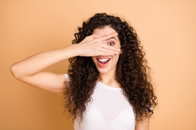 Let's play game! Photo of pretty funny lady hiding eye half playful facial expression fingers wear white casual clothes isolated beige pastel color background