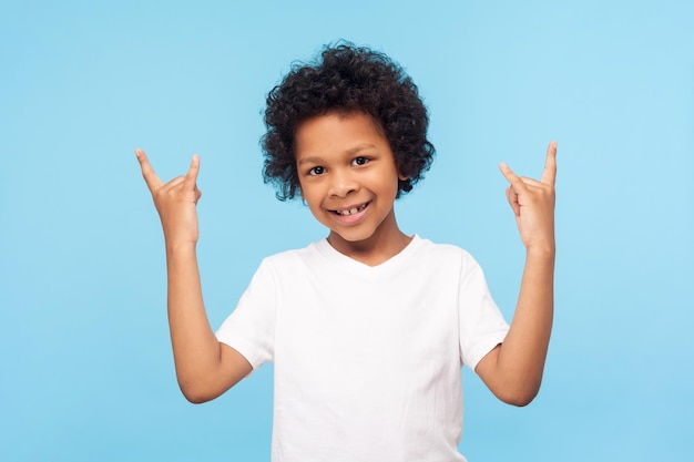 Let39s rock portret van vrolijke funky kleine jongen met krullend haar in witte tshirt tonen rock-'n-roll gebaar en glimlachen maken gek cool symbool met handen studio opname geïsoleerd op blauwe achtergrond