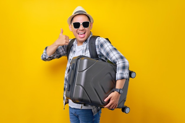 Lets go excited traveler tourist Asian man in casual clothes with hat glasses holding suitcase showing thumb up on yellow background Passenger traveling on weekends Air flight journey concept