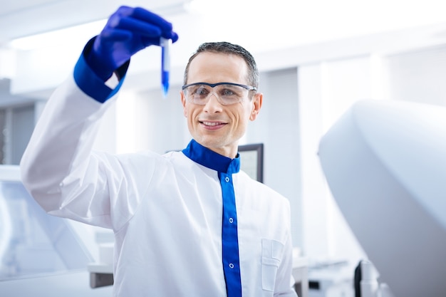 Lets see. Joyfull  determined male researcher jiggling test tube with blue liquid while examining it and standing in the lab