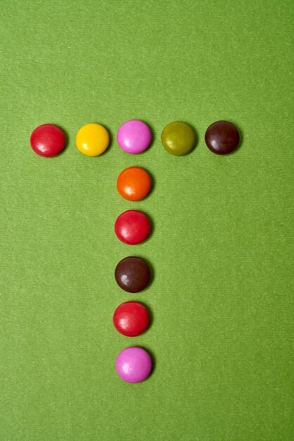 Letter T geschreven met gekleurde ronde chocoladesuikergoed op groene achtergrond.