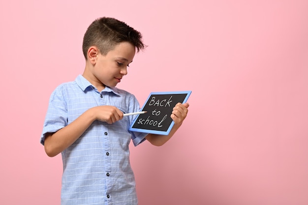 Lettering Back To School on a chalk board in the hands of a happy pupil. Isolated over pink background with copy space