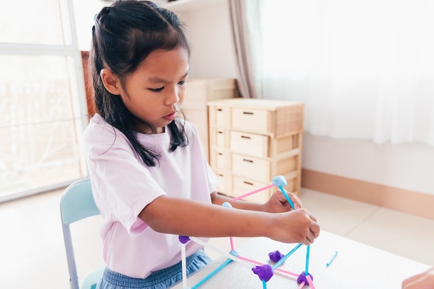 Leuk Aziatisch kindmeisje dat speelt en creëert met speeldeeg en rietjes. Kind geconcentreerd met playdough bouwen van een molecuulmodel. Kid en ambacht met recycle dingen.