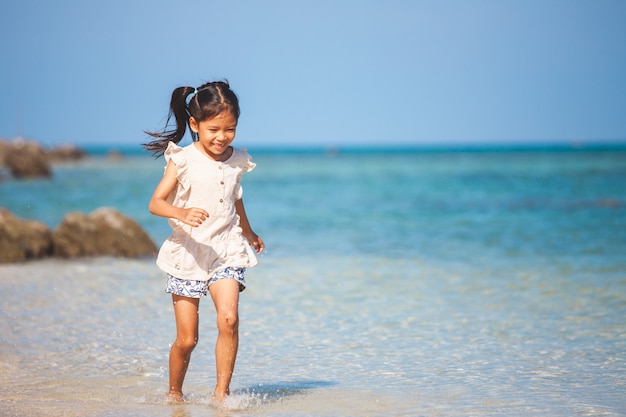 Leuk Aziatisch kindmeisje die pret hebben om op strand dichtbij het mooie overzees in de zomervakantie te spelen en te lopen