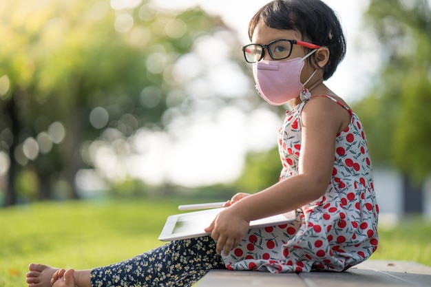 Leuk Aziatisch klein meisje met een gezichtsmasker zittend op het gras en tekenend op een tablet