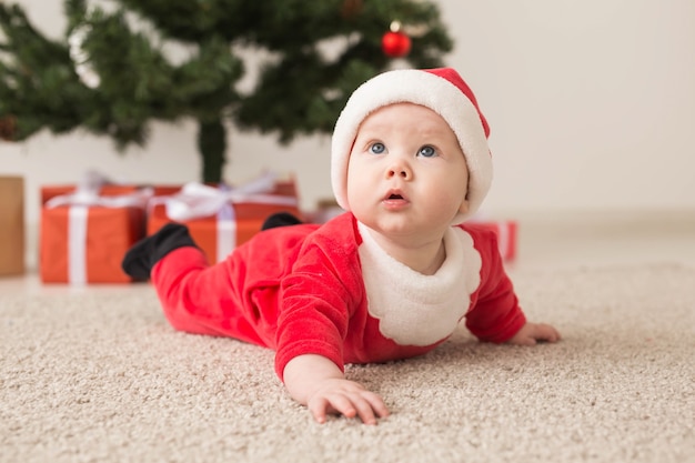 Leuk babymeisje die het kostuum van de Kerstman dragen dat op vloer over Kerstboom kruipt. Vakantie seizoen.