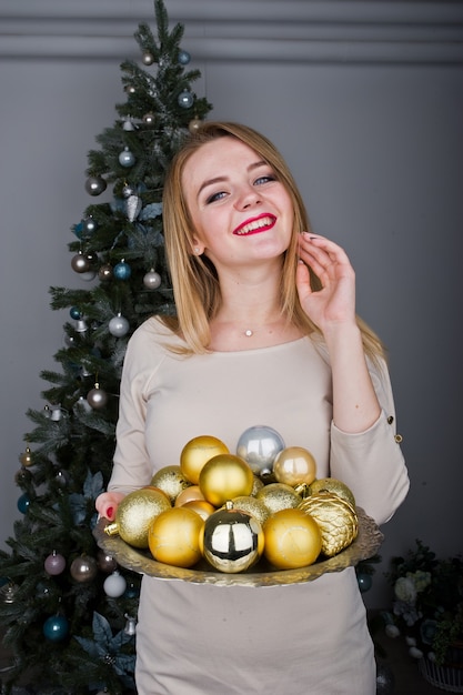 Leuk blond meisje op beige jurk tegen nieuwjaarsboom met gouden nieuwjaarsballen in de studio. Fijne vakantie.