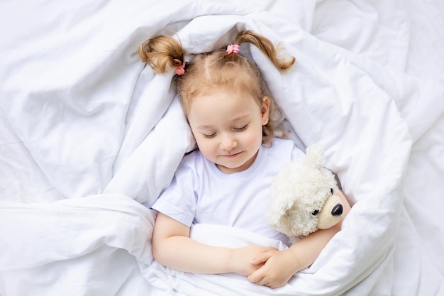 Leuk blond meisje slaapt op een wit katoenen bed thuis en knuffelt een teddybeer.