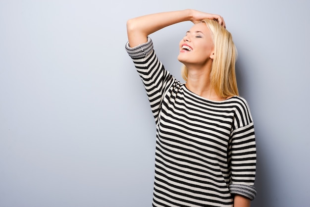 Leuk en flirterig. Mooie jonge blonde haarvrouw die hand in haar houdt en glimlacht terwijl ze tegen een grijze achtergrond staat