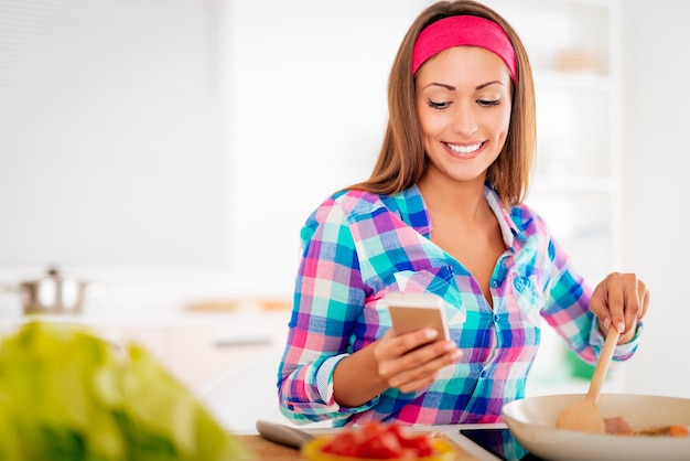 Leuk gelukkig meisje dat een gezonde maaltijd maakt en een bericht leest aan de telefoon in de huishoudelijke keuken. Glimlachen en naar beneden kijken.