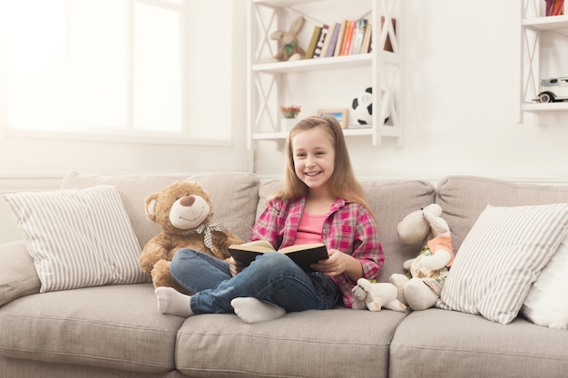 Leuk gelukkig meisje dat teddybeer omhelst en boek leest. Mooi kind thuis, zittend op de bank met haar favoriete speelgoed, plezier hebben, ruimte kopiëren