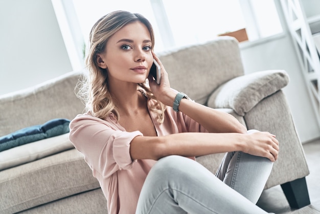 Leuk gesprek. Mooie jonge vrouw die naar de camera kijkt en aan de telefoon praat terwijl ze thuis vloeren