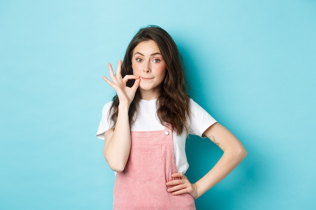 Leuk glamourmeisje dat lippenritsgebaar maakt, haar mond op slot doet, belooft geheim te houden, tegen een blauwe achtergrond staat