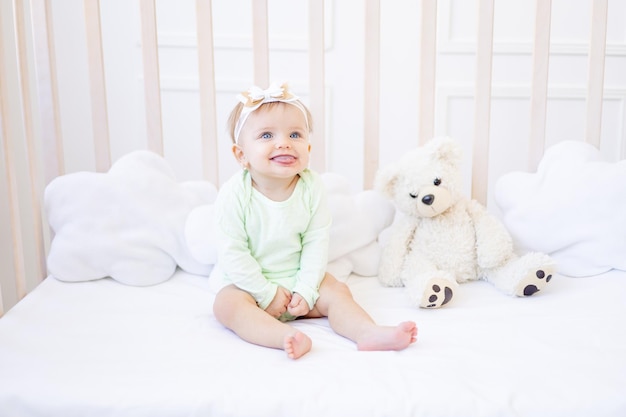 Leuk grappig klein babymeisje toont tong in een wieg op een wit katoenen bed in een groene bodysuit in een lichte kinderkamer en glimlacht