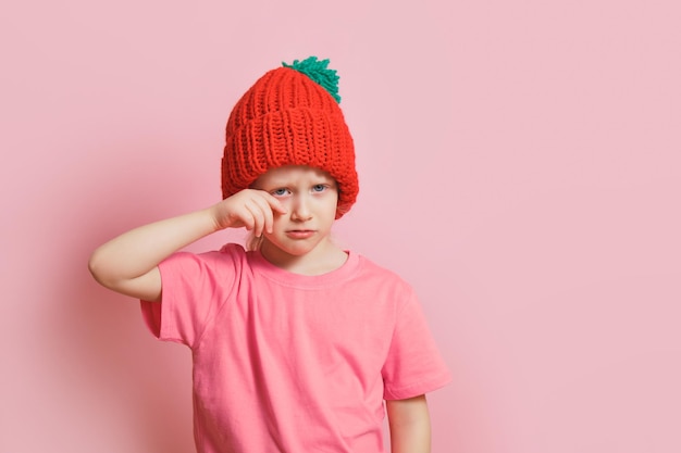 Leuk Kaukasisch peutermeisje in een gebreide muts op een roze achtergrond, een laf kind veegt tranen van haar wangen. emotie van ontevredenheid en teleurstelling