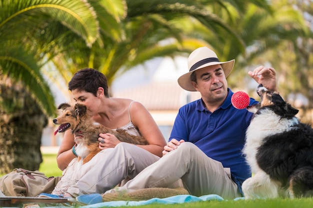Leuk kaukasisch stel van middelbare leeftijd zittend op het gras met palmen op de achtergrond tijdens het spelen met twee grappige en mooie honden en een rode bal, geniet en veel plezier met dierenconcept