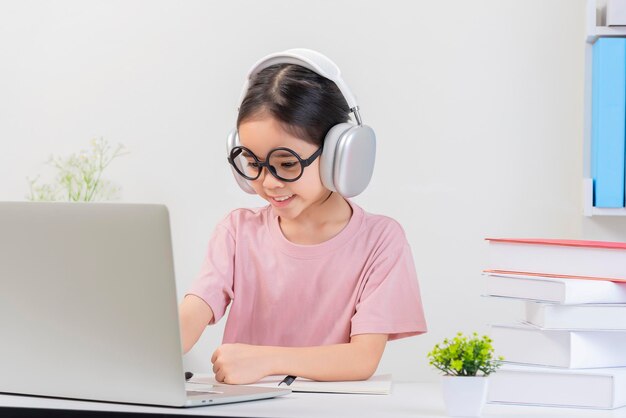 Leuk klein Aziatisch meisje studeert thuis met laptop en draadloze koptelefoon.