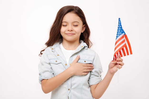 Leuk klein gelukkig meisje dat de Amerikaanse vlag vasthoudt en haar borst aanraakt terwijl ze geluk uitdrukt en met gesloten ogen staat geïsoleerd in een witte muur