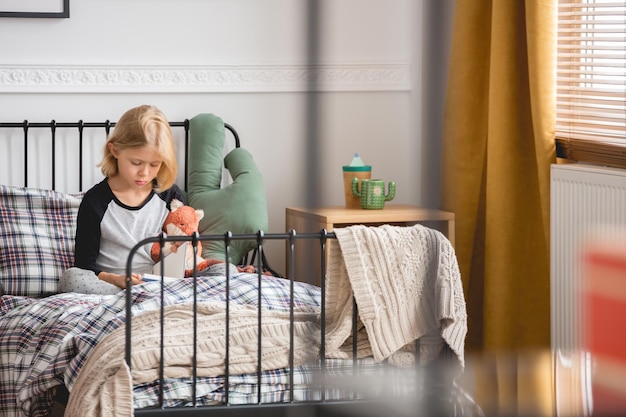 Leuk klein meisje dat op haar enkelvoudige metalen bed zit in een trendy slaapkamerinterieur voor kind