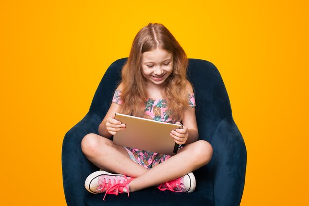 Leuk klein meisje speelt met een tablet zittend in de stoel op een gele muur