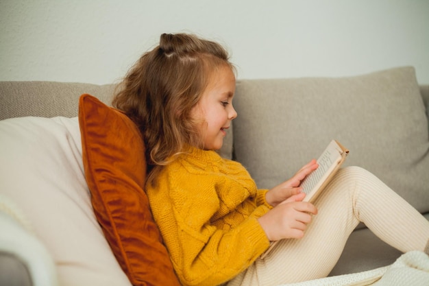 Leuk klein meisje van 4 jaar in een gebreide oranje trui leest een boek in een gezellige kamer.