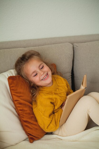 Leuk klein meisje van 4 jaar in een gebreide oranje trui leest een boek in een gezellige kamer.