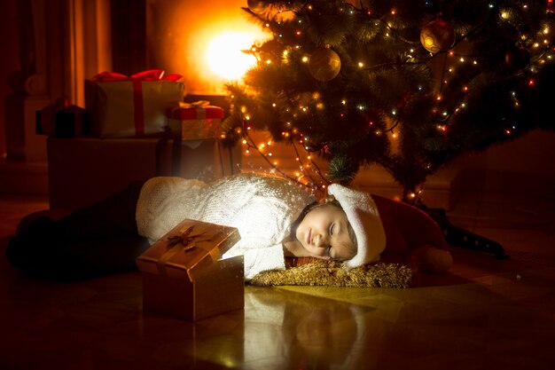 Leuk meisje dat onder de kerstboom ligt en in een gloeiende geschenkdoos kijkt