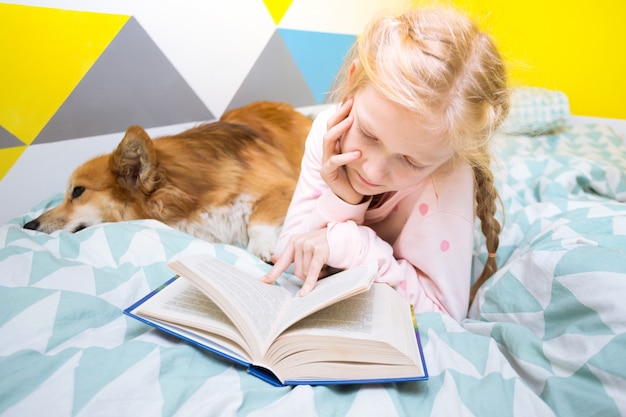Leuk meisje en hond corgi op het bed in de kinderkamer lezen een boek. thuisonderwijs