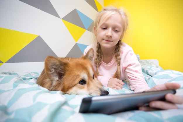 Leuk meisje en hond corgi op het bed in de kinderkamer lezen een boek. thuisonderwijs