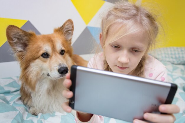 Leuk meisje en hond corgi op het bed in de kinderkamer spelen op de tablet