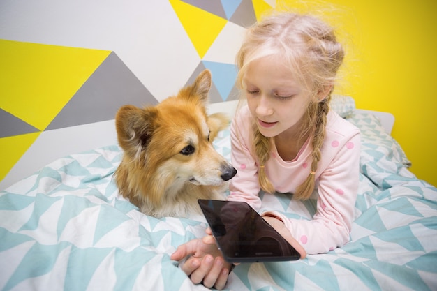 Leuk meisje en hond corgi op het bed in de kinderkamer spelen op de tablet