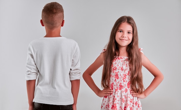 Foto leuk meisje en jongen op een grijze achtergrond. het meisje staat voor de camera, terwijl de jongen met zijn rug naar hem toe staat.