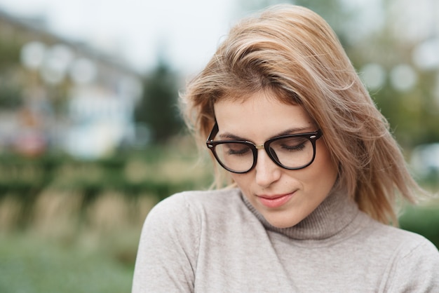 Leuk meisje in een beige trui en een stijlvolle bril, wind fladdert met haar lange blonde haar