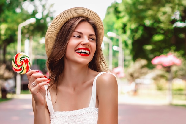 Leuk meisje met rode lippenstift in een strooien hoed en een lolly in haar handen lacht en glimlacht in het park voor tekst. Zomer meisje loopt en heeft plezier in de straten van de stad
