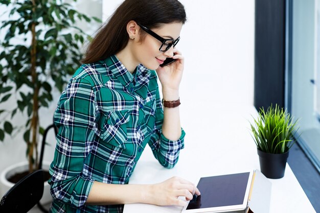 Leuk meisje met zwart haar dragen shirt en bril zitten in café met tablet en kopje koffie, praten op mobiele telefoon, freelance concept.